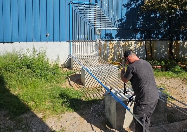 A person in a black shirt and cap works on blue and silver piping by a large blue metal building, sunlight and greenery around.