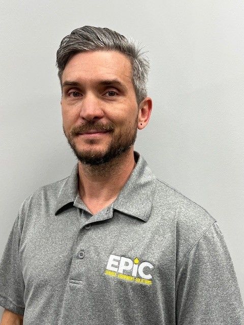 A man with short gray hair and a trimmed beard in a gray EPIC polo stands before a plain light gray background, smiling softly.