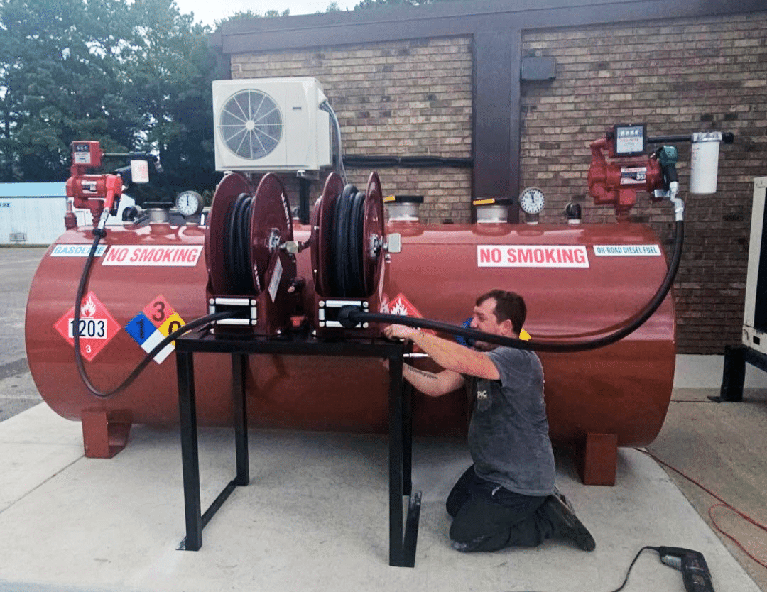 A man kneels while working on hoses attached to a large horizontal fuel tank marked with hazard signs and multiple NO SMOKING warnings outside a brick building.