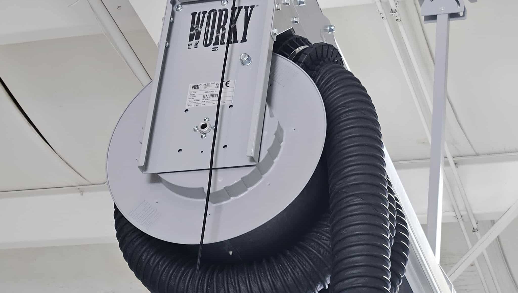 A close-up of a ceiling-mounted industrial hose reel labeled WORKY with a coiled black hose in a bright white-painted workshop ceiling.