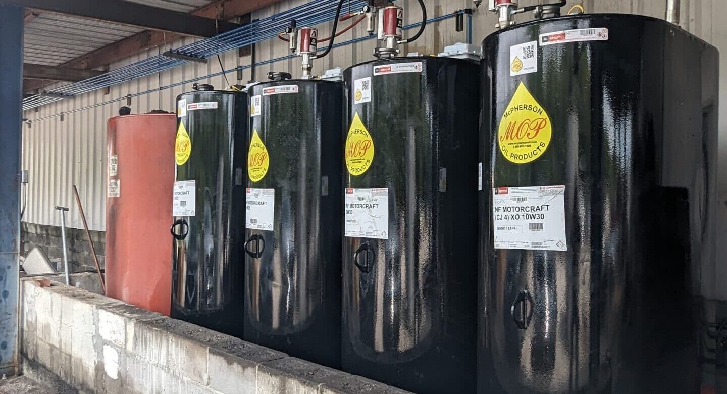 Four black fuel storage tanks with yellow warning labels sit on a concrete platform against a metal wall, pipes and wires above.