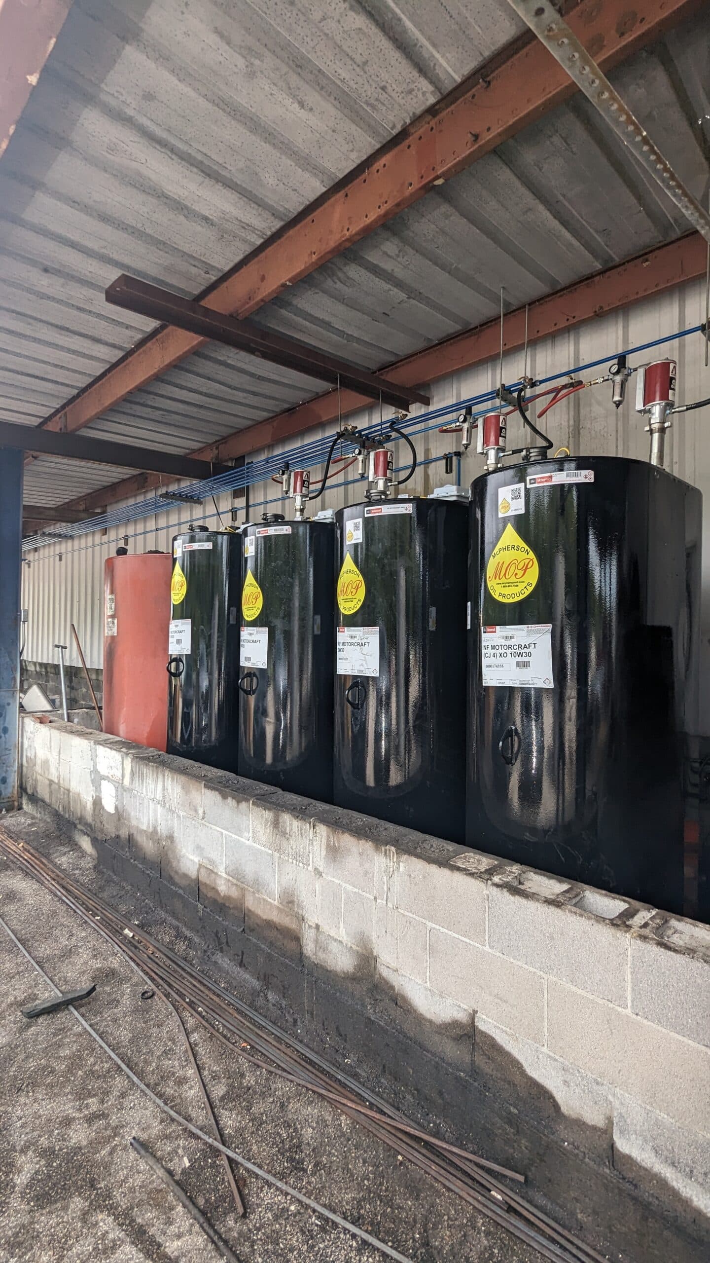 Four large black industrial tanks with yellow safety labels are lined up on a concrete platform, under a metal roof beside a corrugated wall. Pipes and red valves run above the tanks.