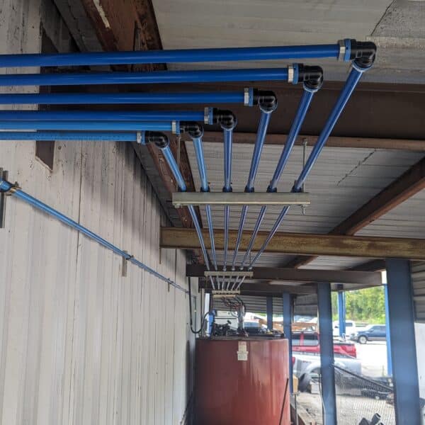 Blue pipes run along a corrugated metal wall under a roof, leading to a red tank in an outdoor area with dirt and parked cars.