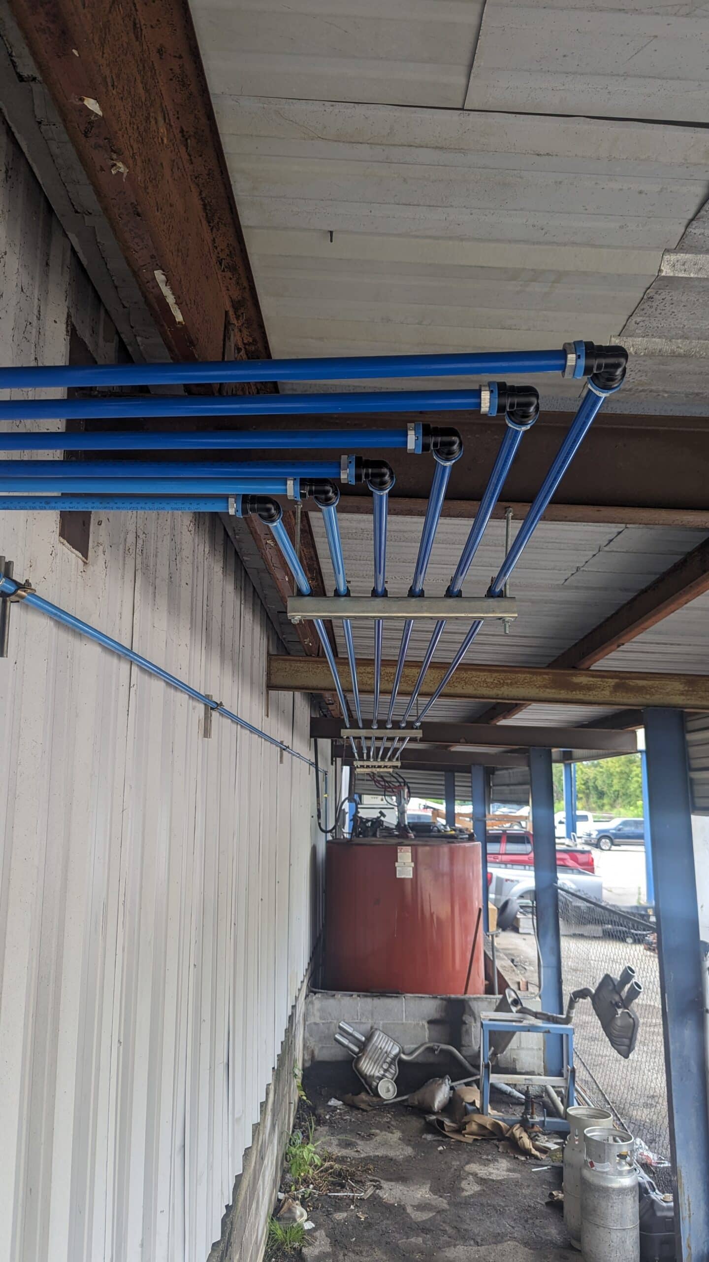 Blue pipes run along a corrugated metal wall under a roof, leading to a red tank in an outdoor area with dirt and parked cars.