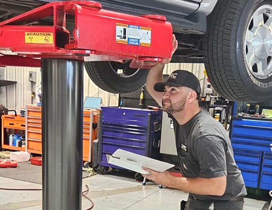 A mechanic checks under a raised car in an auto shop, holding papers. Colorful tool chests and equipment appear in the background.