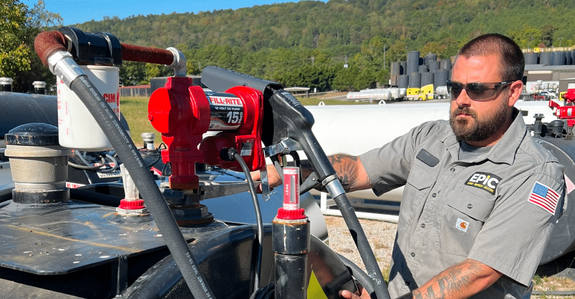 A man wearing sunglasses and a gray EPIC uniform operates industrial fuel equipment outdoors, with large tanks and wooded hills behind.