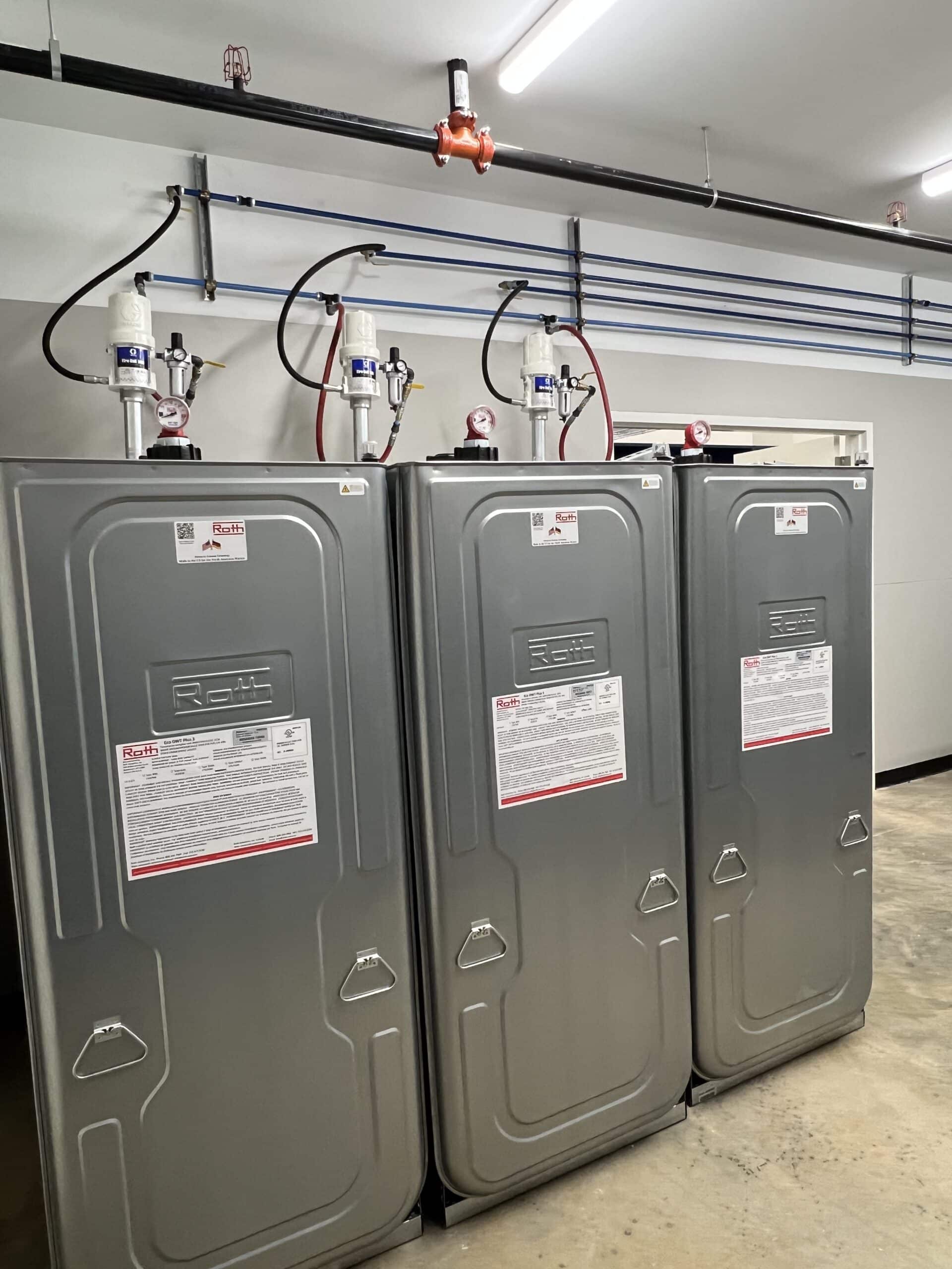 Three large metal tanks with warning labels stand side by side in a bright room, red and black pipes attached to their tops.