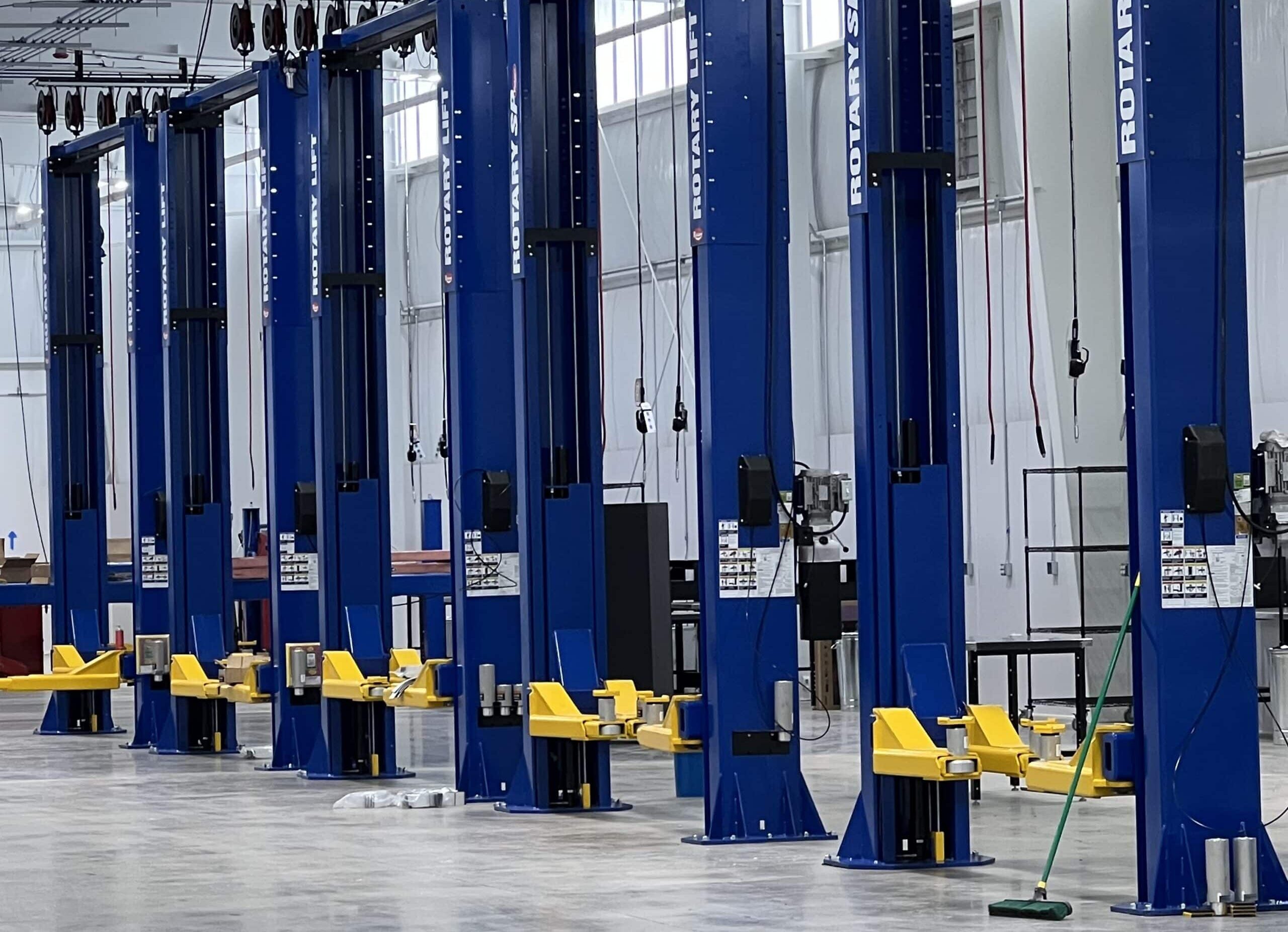 A row of blue vehicle lifts with yellow arms in a bright, spacious garage; cables hang above and a broom lies on the floor out front.