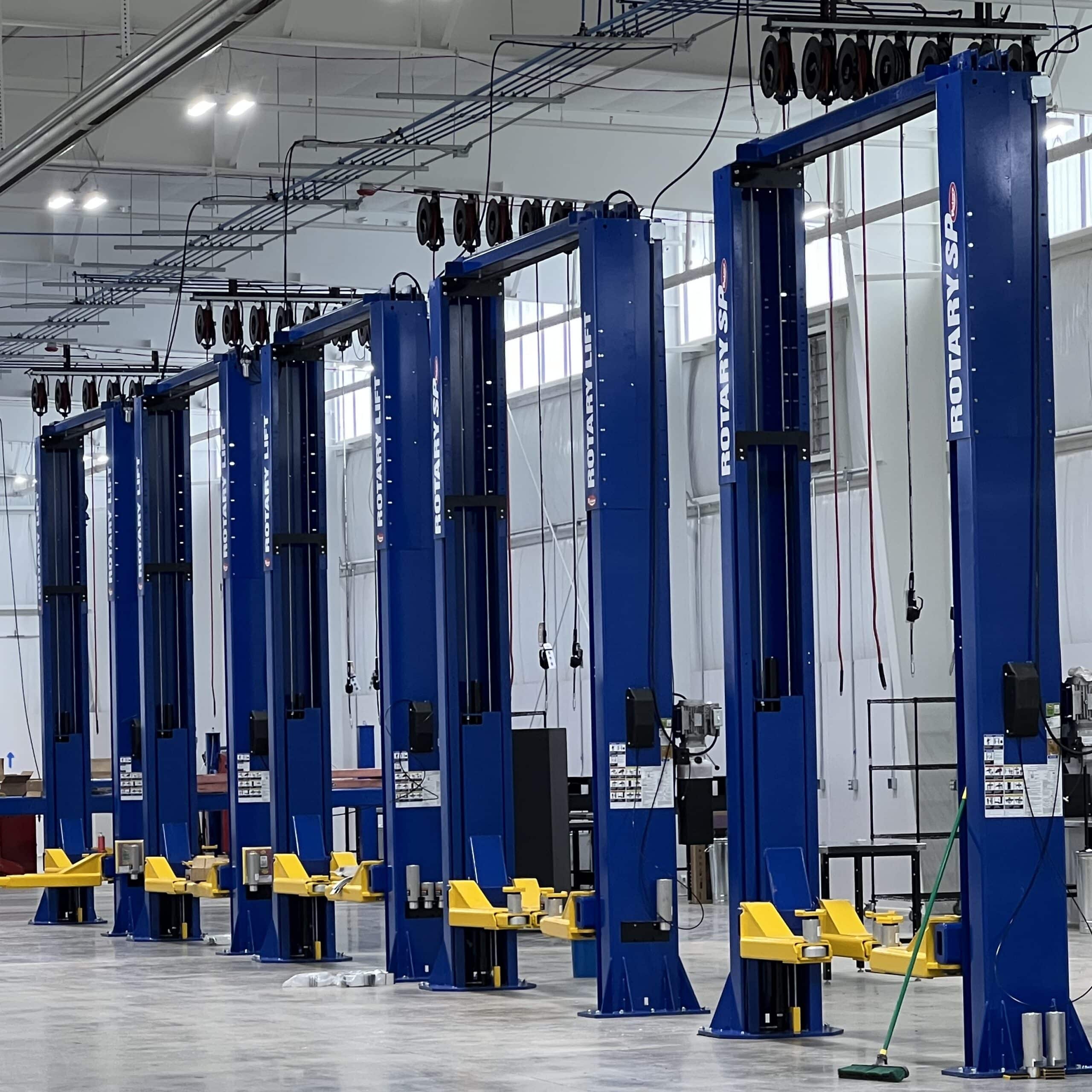 Blue automotive lifts with yellow supports line a spacious, clean garage; cables hang above and cleaning tools sit on the shiny floor.