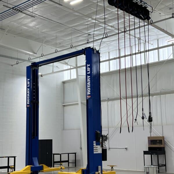 A blue automotive lift in a spacious, clean garage with white walls, high ceiling, black tables, and cables hanging overhead.