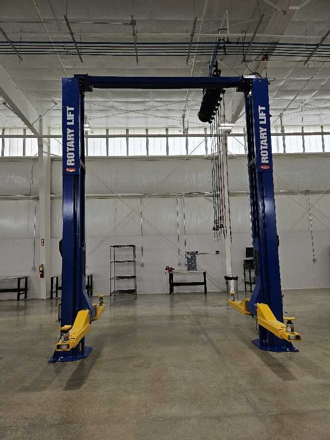 A blue Rotary Lift car lift in an empty, brightly lit garage with white walls, concrete floors, shelves, and tools in the background.