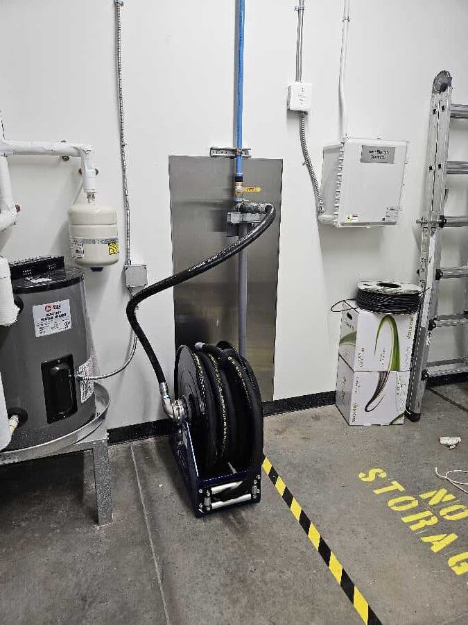 A hose reel on the floor of a utility room with pipes, electrical boxes, a ladder, and a NO STORAGE zone marked by yellow tape.
