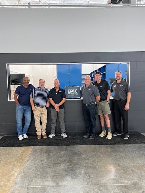 Six men stand in a row smiling in front of a black and gray wall. One holds a plaque that says EPIC. They are indoors.