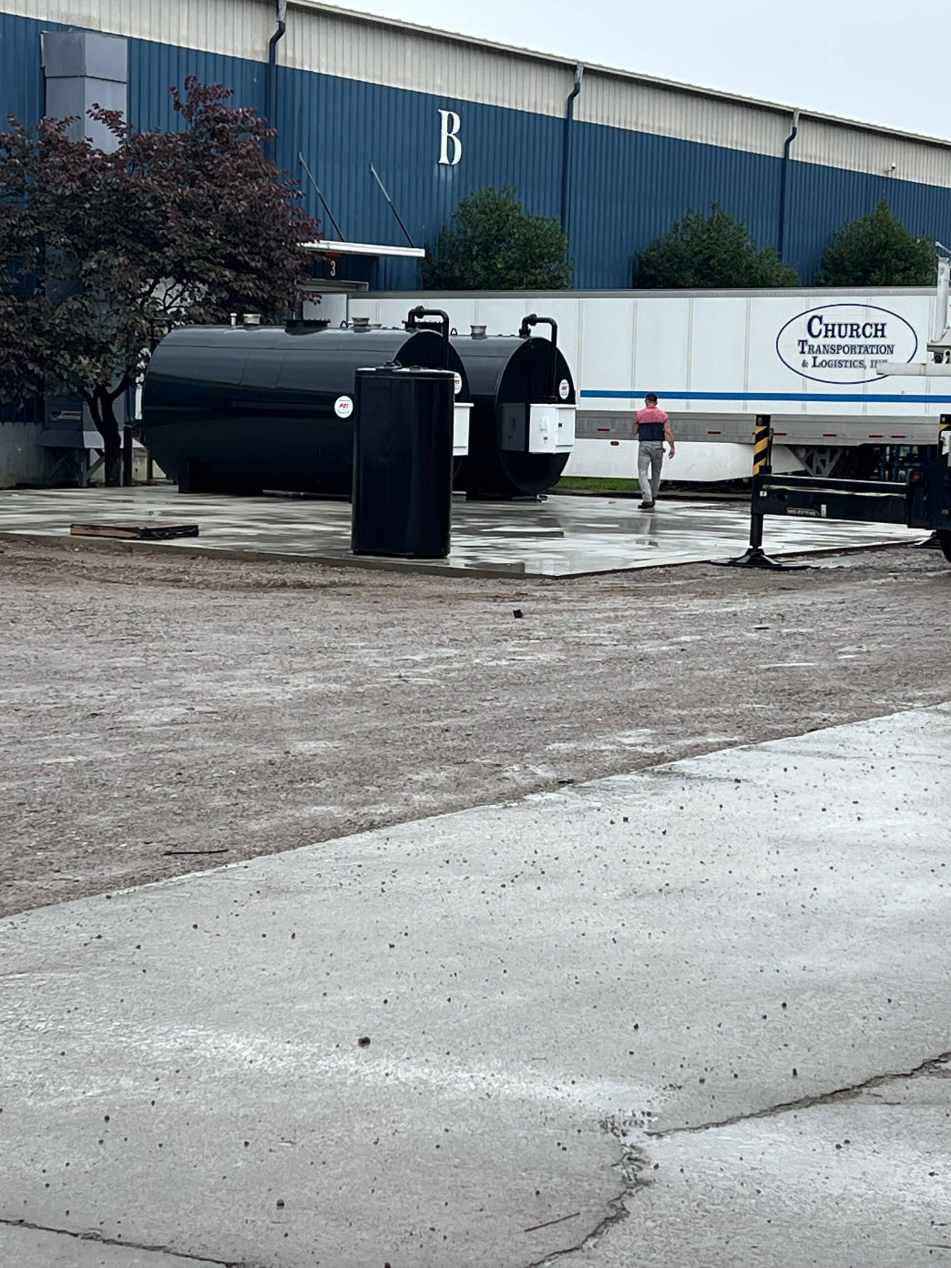 A person walks near large black and white industrial tanks and a Church Transportation & Logistics truck trailer outside a blue warehouse with the letter B on it, on a cloudy day.