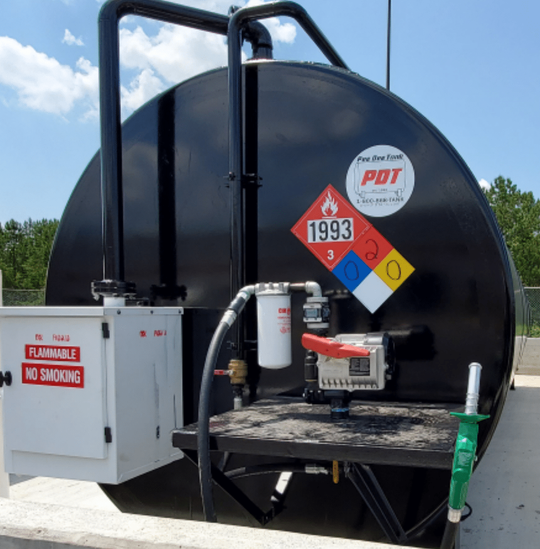 A large black fuel storage tank with hazard labels, a pump, attached hoses, and warning signs for FLAMMABLE and NO SMOKING outdoors.